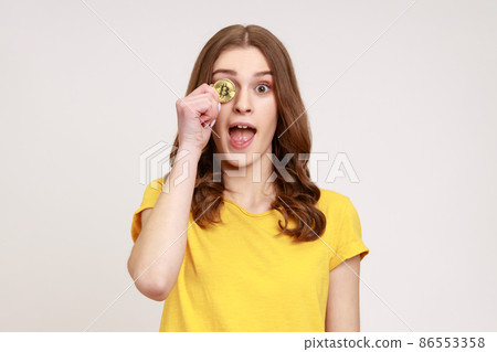 Portrait of playful excited woman of young age with charming smile wearing yellow T-shirt covering one eye with golden bitcoin, cryptocurrency. Indoor studio shot isolated on gray background. 86553358