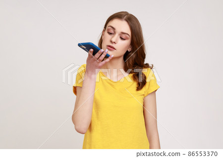 Innovative technologies. Young attractive woman with brown wavy hair asking voice assistant on cell phone, giving task, recording message. Indoor studio shot isolated on gray background. Innovative technologies. Young attractive woman with brown wavy hair asking voice assistant on cell phone, giving task, recording message. Indoor studio shot isolated on gray background. 86553370