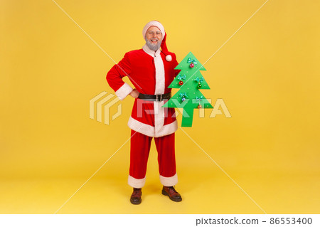 Confident adult man wearing santa claus costume standing with hand on hip, holding paper green Christmas tree, looks at camera with smile. Indoor studio shot isolated on yellow background. 86553400