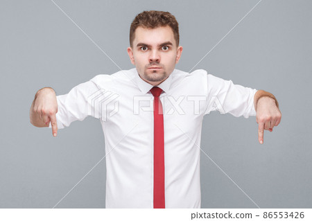 Here and right now. Portrait of young serious handsome man in white shirt and tie standing and looking at camera with fingers down. indoor isolated on gray background. Here and right now. Portrait of young serious handsome man in white shirt and tie standing and looking at camera with fingers down. indoor isolated on gray background. 86553426