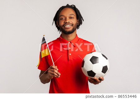 Man with toothy smile wearing red casual style T-shirt, holding flag of germany and soccer black and white classic ball and watching match. Indoor studio shot isolated on gray background. 86553446