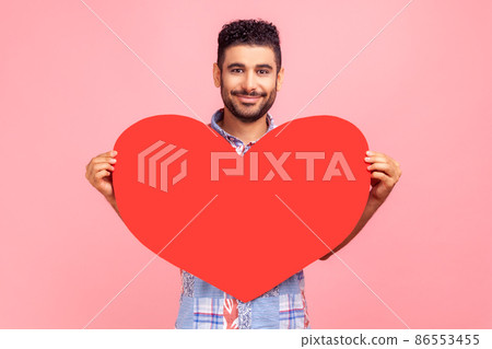 Portrait of smiling bearded man wearing blue shirt holding big red heart in hands, looking at camera with positive, expressing love. Indoor studio shirt isolated on pink background. Portrait of smiling bearded man wearing blue shirt holding big red heart in hands, looking at camera with positive, expressing love. Indoor studio shirt isolated on pink background. 86553455