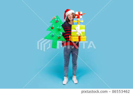 Full length portrait of woman with wavy hair in casual sweater and santa claus hat, holding decorative tree and present boxes, celebrating Christmas. Indoor studio shot isolated on blue background. 86553462