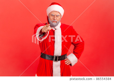 Elderly man with gray beard in santa claus costume pointing at camera with angry expression and accusing blaming, indicating at you, making choice. Indoor studio shot isolated on red background. 86554384