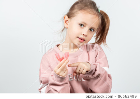 Cute little girl with blond hair is holding a pink dental myofunctional trainer Cute little girl with blond hair is holding a pink dental myofunctional trainer 86562499