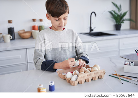 Cheerful concentrated child boy smiling and painting Easter egg at the kitchen Cheerful concentrated child boy smiling and painting Easter egg at the kitchen 86565644