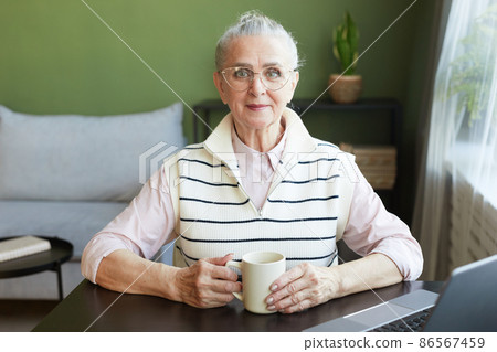 Happy mature female in eyeglasses and casual clothes having tea by table Happy mature female in eyeglasses and casual clothes having tea by table 86567459