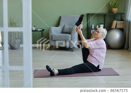 Senior woman in activewear doing physical exercise on yoga mat in living-room Senior woman in activewear doing physical exercise on yoga mat in living-room 86567575
