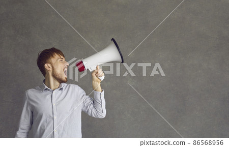 Young determined man makes a loud announcement while speaking into a loudspeaker. Skinny red-haired man in a shirt stands on a gray background near the free space for text. Advertising concept. Young determined man makes a loud announcement while speaking into a loudspeaker. Skinny red-haired man in a shirt stands on a gray background near the free space for text. Advertising concept. 86568956