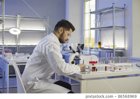 Millennial Caucasian man laboratory assistant in white medical uniform make research experiments in lab. Young technician or tester use microscope, test tube glass do chemistry science investigation. Millennial Caucasian man laboratory assistant in white medical uniform make research experiments in lab. Young technician or tester use microscope, test tube glass do chemistry science investigation. 86568969