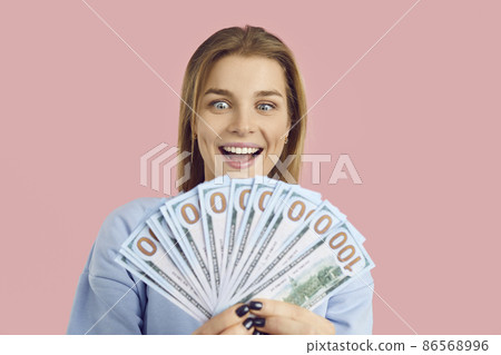 Wealthy woman isolated on pink background smiling and holding bunch of paper banknotes in her hands. Young girl who wins lots of money and becomes rich looking at prize cash with happy face expression 86568996