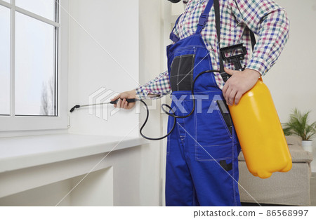 Pest control guy spraying poisonous gas or liquid on a windowsill at home. Male exterminator in overall uniform workwear spraying insecticide over a modern white window and sill inside the house 86568997