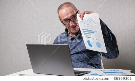 Elderly man checks drawings near laptop at white office desk 86569361