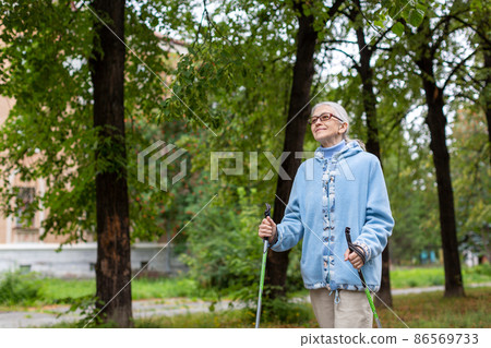 Mature woman active lifestyle. Caucasian old lady in blue hoodie and glasses does nordic walking in the park outdoor in late summer, selective focus. Mature woman active lifestyle. Caucasian old lady in blue hoodie and glasses does nordic walking in the park outdoor in late summer, selective focus. 86569733