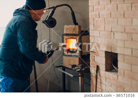 Blacksmith forge oven with hot flame. Smith put in and heating iron piece of steel in fire of red hot forge oven Blacksmith forge oven with hot flame. Smith put in and heating iron piece of steel in fire of red hot forge oven 86571016