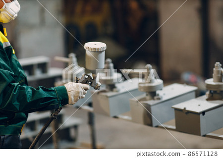 Powder primer coating of metal parts. Worker man in a protective suit sprays powder paint from gun on metal product construction at factory plant. Powder primer coating of metal parts. Worker man in a protective suit sprays powder paint from gun on metal product construction at factory plant. 86571128