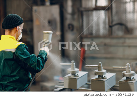 Powder primer coating of metal parts. Worker man in a protective suit sprays powder paint from gun on metal product construction at factory plant. 86571129