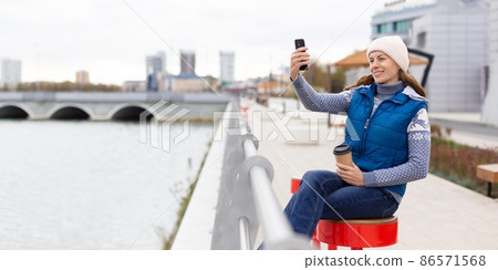 Video call. Adult smiling woman in knit cap sits with coffee on the city embankment and using her mobile, format photo 1x2, selected focus. 86571568