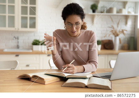 Serious young Latina woman studying seated at dining table Serious young Latina woman studying seated at dining table 86572275