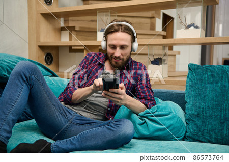 Caucasian young man using social media at home. Handsome guy with wireless headphones relaxing on the couch at home Caucasian young man using social media at home. Handsome guy with wireless headphones relaxing on the couch at home 86573764