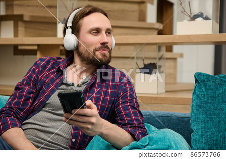 Middle aged Caucasian bearded handsome man with wireless headphones, holds a smartphone in his hand and dreamily looking aside 86573766