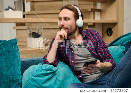 Middle aged bearded handsome European man with wireless headphones, holds a smartphone in his hand and dreamily looking aside, enjoying day off, relaxing on a sofa at home environment 86573767