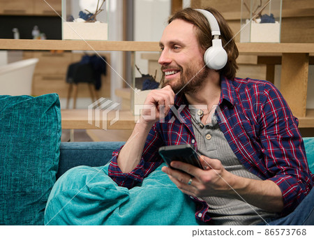 Charming handsome attractive Caucasian bearded man in casual attires holding mobile phone and smiling with cheerful toothy smile looking aside while listening music with wireless headphones at home 86573768