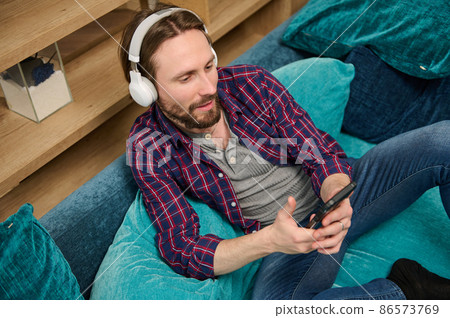 Top view of a middle aged bearded handsome Caucasian man with wireless headphones, holds a smartphone in his hand and dreamily looking aside, enjoying day off, relaxing on a sofa at home environment Top view of a middle aged bearded handsome Caucasian man with wireless headphones, holds a smartphone in his hand and dreamily looking aside, enjoying day off, relaxing on a sofa at home environment 86573769