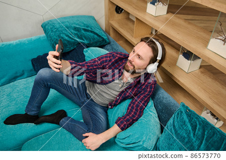 Overhead view of handsome relaxed bearded Caucasian man wearing wireless headphones, talking by video call, looking at web camera on the smartphone in his hand, relaxing lying down on a sofa at home Overhead view of handsome relaxed bearded Caucasian man wearing wireless headphones, talking by video call, looking at web camera on the smartphone in his hand, relaxing lying down on a sofa at home 86573770