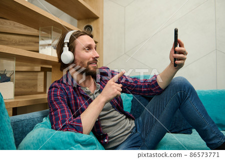 Handsome relaxed bearded Caucasian man wearing wireless headphones, talking by video call, looking at web camera on the smartphone in his hand, relaxing lying down on a sofa at home Handsome relaxed bearded Caucasian man wearing wireless headphones, talking by video call, looking at web camera on the smartphone in his hand, relaxing lying down on a sofa at home 86573771