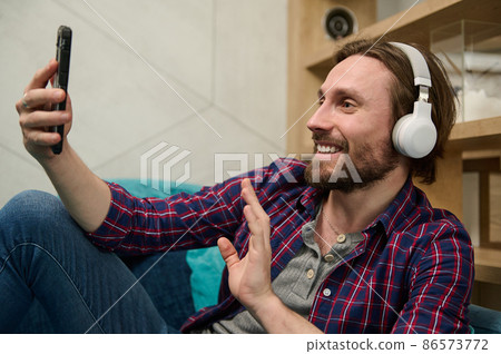 Handsome man having fun discussion by video call, wearing wireless headphones and smiles looking at a smartphone web camera, greeting and waving hello while relaxing at home 86573772