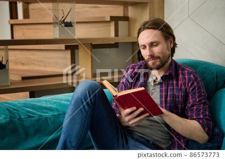 Portrait of a handsome charming bearded middle aged Caucasian man reading book, sitting on sofa at home interior, enjoying a weekend in calm environment 86573773