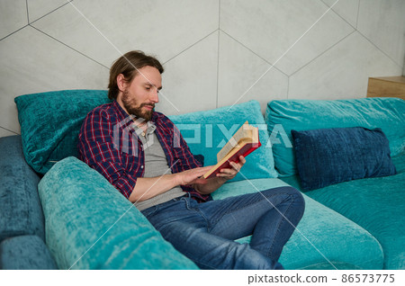 Serene confident young bearded Caucasian man relaxing on the couch, reading book in home environment. Education, study, knowledge and erudition concept 86573775