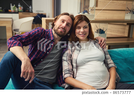 Portrait of a happy young heterosexual loving couple in anticipation of a baby relaxing together on the sofa at home, smiling looking at camera. Beautiful pregnant woman, carefree pregnancy concept 86573777