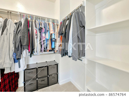 Small walk-in closet with hanging sweaters, shirts and blouse on the metal rods 86574764
