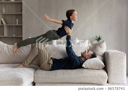 Happy father lying on couch, lifting daughter pretending flying Happy father lying on couch, lifting daughter pretending flying 86578741