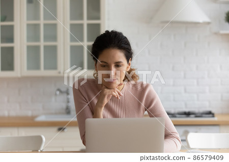 Hispanic woman sit in kitchen web surfing using laptop 86579409