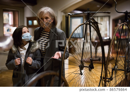 Elderly woman with preteen granddaughter in protective masks examining retro bicycles in museum 86579864