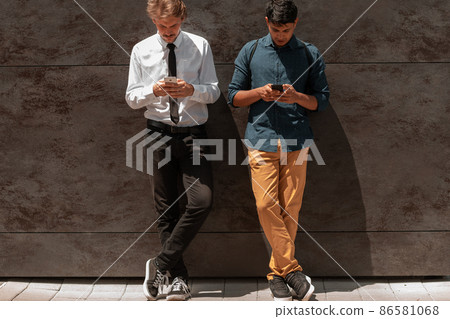 Casual multiethnic startup business men one of them is Indian using smartphone during break from work in front of grey wall outside Casual multiethnic startup business men one of them is Indian using smartphone during break from work in front of grey wall outside 86581068