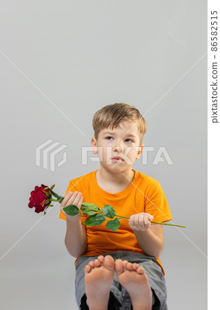 adorable preschool sitting barefoot. He is looking at camera while holding rose. On grey background. 86582515