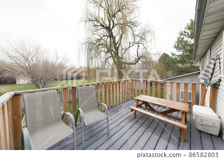 Deck of a house with gray armchairs, outdoor dining table with bench, cooler container and umbrella 86582803