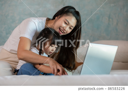 Beautiful Asian mom teaching her daughter to learn and using computer laptop for preschool or watching cartoons online at home.Happy mother relax with little girl having fun with laptop at home 86583226