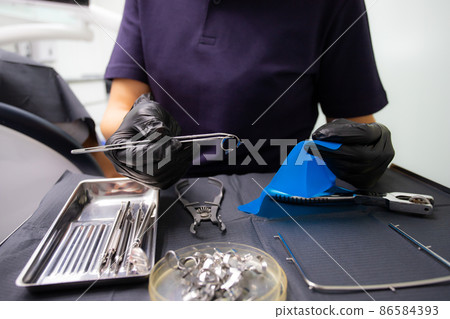 A dentist prepares a latex scarf with metal clips to treat canals and pulpitis. Pliers punch a hole in a black latex insulating napkin A dentist prepares a latex scarf with metal clips to treat canals and pulpitis. Pliers punch a hole in a black latex insulating napkin 86584393