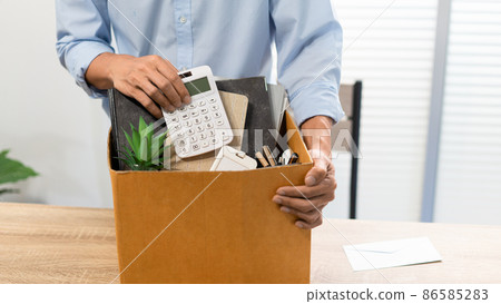 Resignation Concept The man in light blue shirt standing at the desk and putting the calculator and other stuff into the box Resignation Concept The man in light blue shirt standing at the desk and putting the calculator and other stuff into the box 86585283