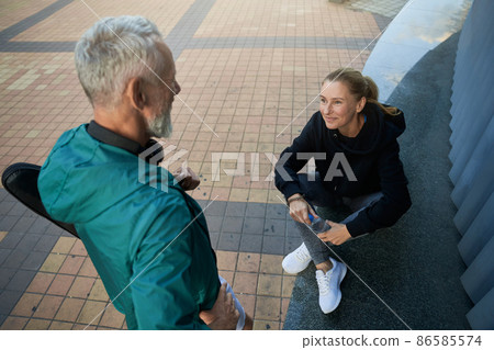 High angle view of sportive mature couple, man and woman in sportswear talking outdoors while waiting together for workout High angle view of sportive mature couple, man and woman in sportswear talking outdoors while waiting together for workout 86585574