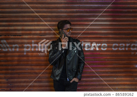 Portrait of young stylish brunet caucasian man in black leather jacket and yellow sunglasses. Standing near the wall and talking by phone. 86589162