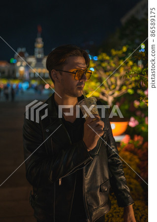 Portrait of young handsome brunet caucasian man in black leather jacket and yellow sunglasses. Standing on the street at night and drinking ice coffee 86589175