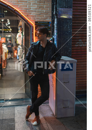 Portrait of young stylish brunet caucasian man in black leather jacket and yellow sunglasses. Standing on the street near the wall in evening.  86589321