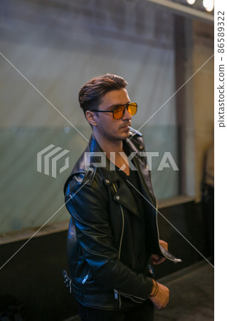 Portrait of young handsome brunet caucasian man in black leather jacket and yellow sunglasses. Holding cigarette in his mouth. High quality photo 86589322