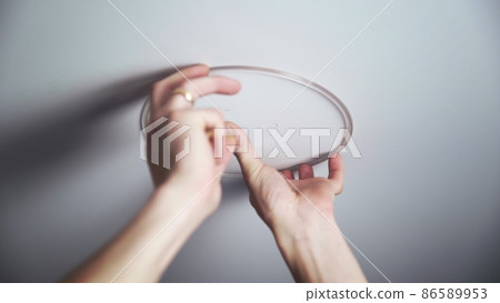 A close-up of the hands of an unrecognizable man setting up the base for mounting a ceiling light. Repair A close-up of the hands of an unrecognizable man setting up the base for mounting a ceiling light. Repair 86589953
